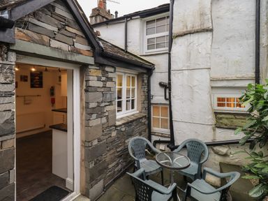 An outdoor patio area with a glass table and four plastic chairs next to a stone and white plaster building at Birchcroft in Ambleside