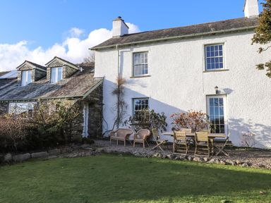 A white house with wooden outdoor chairs and table on a gravel area and a green lawn in front at Robin Cottage in Troutbeck