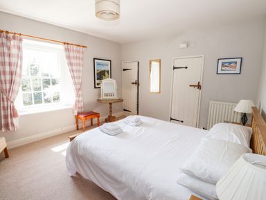 A bedroom with a large bed neatly made with white linens a bedside table with a lamp a window with pink checkered curtains a small wooden table with a mirror and a stool at Robin Cottage in Troutbeck