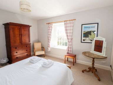 A bedroom with a bed with towels on it a wooden wardrobe an armchair a table with a mirror and a window with checkered curtains at Robin Cottage in Troutbeck