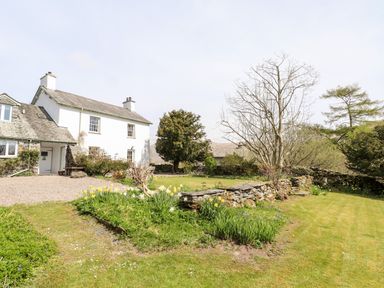 An exterior view of a white cottage with a gravel driveway trees stone walls and garden beds with plants and flowers at Robin Cottage in Troutbeck
