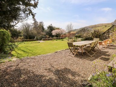 An outdoor garden area with gravel and wooden table and chairs surrounded by grassy lawn and stone walls at Robin Cottage in Troutbeck