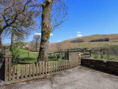 A wooden fence and gate around a garden with outdoor chairs and a table near trees and hills at Boxwood Cottage in Troutbeck