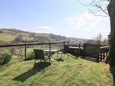 Two chairs and a small round table on grass with wooden fence and gate overlooking green hills at Boxwood Cottage in Troutbeck
