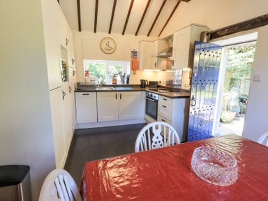 A kitchen with white cabinets a clock on the wall a red tablecloth on a dining table and a blue door open to the outside at Stablemans Cottage at Stepping Stones Ambleside