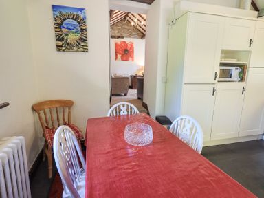 A dining area with a table covered by a red tablecloth surrounded by white chairs and a wooden chair next to a radiator and a white cabinet with a microwave at Stablemans Cottage at Stepping Stones Ambleside