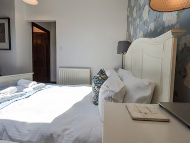 A bedroom with a white bed and pillows next to a nightstand with a lamp and decorative wallpaper at Stablemans Cottage at Stepping Stones Ambleside
