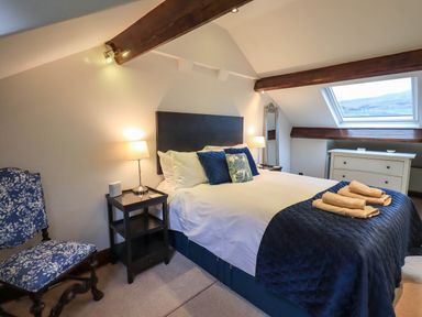 A bedroom with a bed with blue and white bedding a chair a dresser and a window on a slanted ceiling at Stablemans Cottage at Stepping Stones in Ambleside