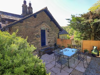 Outdoor patio with metal table and chairs beside stone cottage with blue door and greenery at Stablemans Cottage at Stepping Stones in Ambleside
