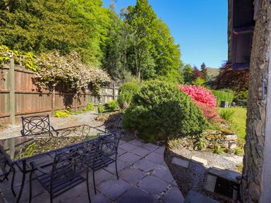 An outdoor patio with metal table and chairs next to a garden with bushes and flowers at Stablemans Cottage at Stepping Stones in Ambleside