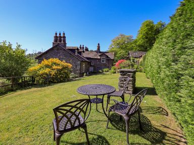 A garden area with a round metal table and four chairs near stone buildings and tall green hedges at Stablemans Cottage at Stepping Stones in Ambleside