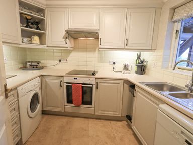 A kitchen with white cabinets a washing machine an oven with red oven mitts a kettle a toaster and a sink by a window at Ghyll Heights in Ambleside