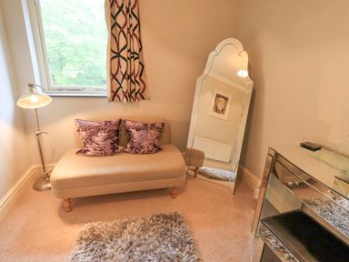A small sitting area with a beige loveseat with two purple pillows a floor lamp a standing mirror a window with patterned curtains a mirrored desk and a shaggy rug at Ghyll Heights in Ambleside