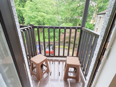 A small balcony with two brown stools and black railing overlooking trees and parked cars at Ghyll Heights Ambleside