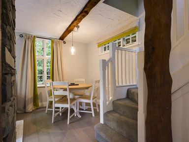 A dining area with a round wooden table and four chairs next to a window and carpeted stairs at Acorn Cottage in Grasmere