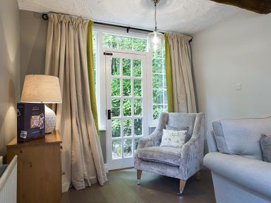 A sitting area with a cushioned armchair a sofa a wooden cabinet with a lamp and glass door with curtains at Acorn Cottage in Grasmere