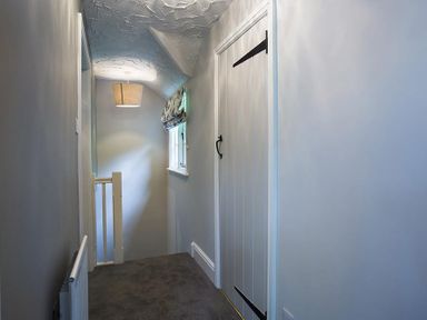 A narrow hallway with a carpeted floor a window with a patterned blind a ceiling light and a white door with black hinges at Acorn Cottage in Grasmere