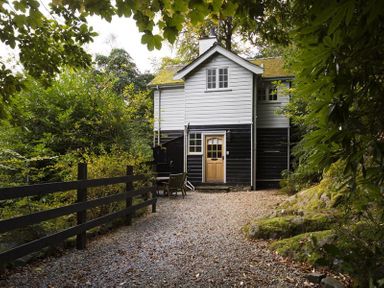 A two-story house with black and white siding and a wooden door surrounded by trees and a gravel pathway at Acorn Cottage in Grasmere