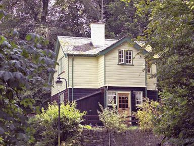 A two-story house with cream and dark brown exterior surrounded by trees and shrubs at Acorn Cottage in Grasmere