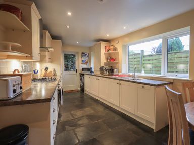 A long kitchen with white cabinets a brown countertop a sink a gas stove a toaster and a small round dining table with chairs at Wainwright Cottage in Ambleside