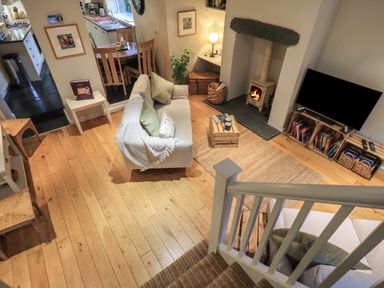 A living room with a fireplace a TV on a wooden stand sofas with cushions a wooden floor and a dining area in the background at Wainwright Cottage in Ambleside