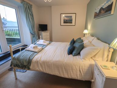 A bedroom with a double bed with white bedding and blue cushions a small white dresser with a tv and lamps on bedside tables at Wainwright Cottage in Ambleside