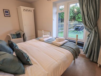 A bedroom with a bed with folded towels pillows and blankets a white wardrobe a wicker chair and a glass door overlooking a garden at Wainwright Cottage in Ambleside