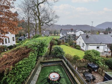 A backyard with a patio table and chairs surrounded by hedges and houses in the background at Wainwright Cottage in Ambleside
