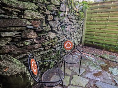 An outdoor stone patio area with two metal chairs with circular orange and white patterned backs against a stone wall and wooden fence at Wainwright Cottage in Ambleside