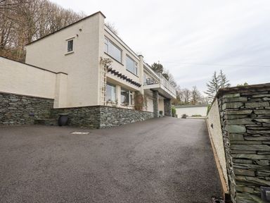 A driveway leading up to a two-story house with stone and white exterior walls with trees in the background at Tanna Hill in Bowness