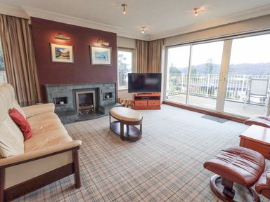 A living room with a sofa, armchair, coffee table, fireplace, television, and large sliding glass doors at Tanna Hill in Bowness