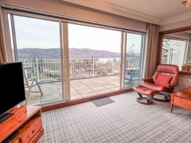 A room with a large sliding glass door opening to a balcony with chairs and a view of hills and trees at Tanna Hill in Bowness