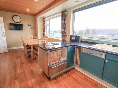 A kitchen and dining area with wooden table and chairs blue cabinets and large windows showing a tree view at Tanna Hill in Bowness