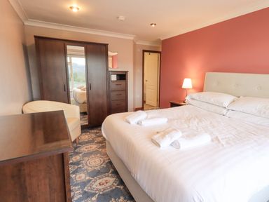 A bedroom with a large bed with white linens and towels on it a bedside table with a lamp a wardrobe with a mirror a chest of drawers an armchair and a door at Tanna Hill in Bowness