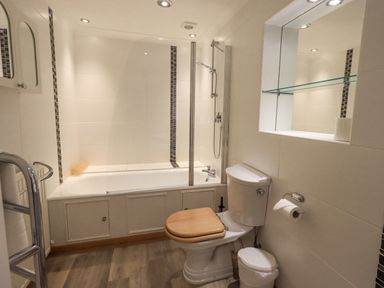 A bathroom with a bathtub and shower, a toilet with a wooden seat, a towel rail with a towel, and a recessed shelf with a glass shelf at Tanna Hill in Bowness