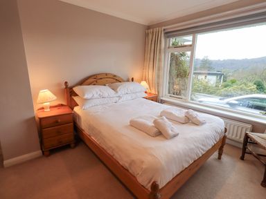 A bedroom with a double bed pillows folded towels bedside tables lamps and a large window overlooking a garden and parked car at Tanna Hill in Bowness
