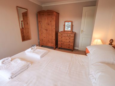 A bedroom with a bed, wooden wardrobe, chest of drawers with a mirror, a wall mirror, and a lamp at Tanna Hill in Bowness