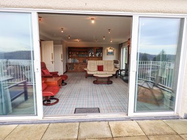 A living room with leather sofas and armchairs and a wooden cabinet visible through sliding glass doors at Tanna Hill in Bowness