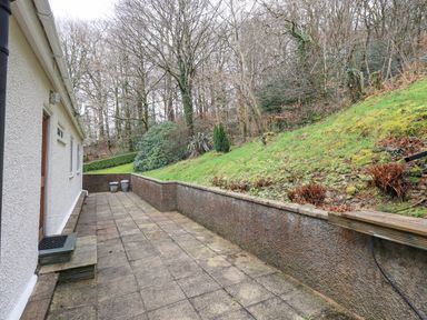 A paved patio area beside a white building with a sloped grassy hill and bare trees in the background at Tanna Hill in Bowness