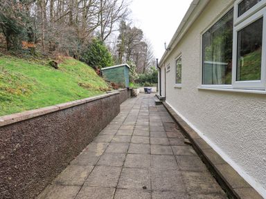 An outdoor paved pathway beside a building with windows a low stone wall and a hillside with trees and a small shed at Tanna Hill in Bowness