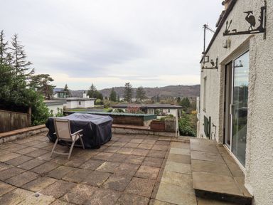A patio area with a covered table and chair on tiled flooring overlooking houses and trees at Tanna Hill in Bowness