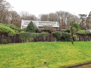 A house with a balcony behind a sloped grassy yard and wooden fence with bushes at Tanna Hill in Bowness