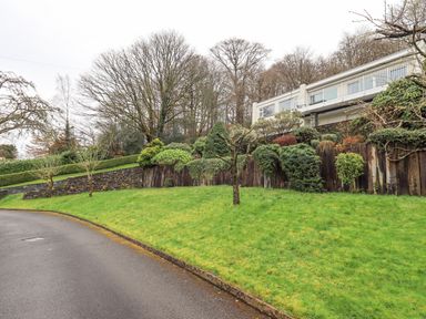 A sloped garden with grass shrubs and trees next to a paved road and a house with a balcony in the background at Tanna Hill in Bowness