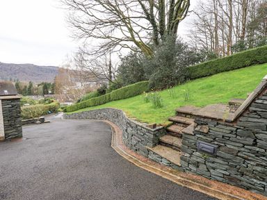 A driveway with stone retaining walls and steps leading to a grassy area with trees at Tanna Hill in Bowness