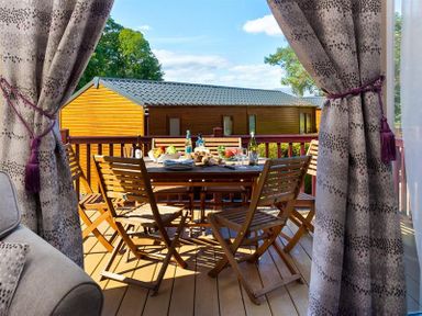 An outdoor wooden dining table with chairs on a deck with food and drinks visible through open curtains at Lancashire Rose in Ambleside