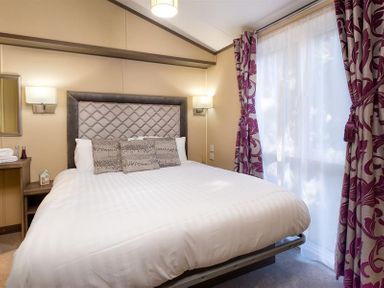 A bedroom with a bed with a padded headboard, three pillows, two wall lamps, a small table with towels and decorative items, and a window with sheer curtains and patterned drapes at Lancashire Rose in Ambleside