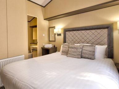 A bedroom with a bed a headboard three pillows bedside tables a lamp and an open door to a bathroom at Lancashire Rose Ambleside
