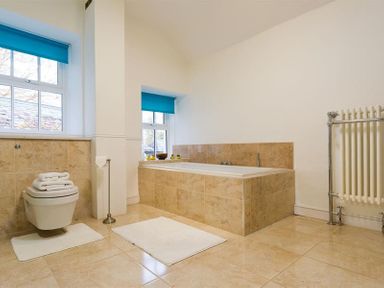 A bathroom with a bathtub tiled in beige and a wall-mounted toilet under windows with blue blinds at High Cleabarrow in Windermere