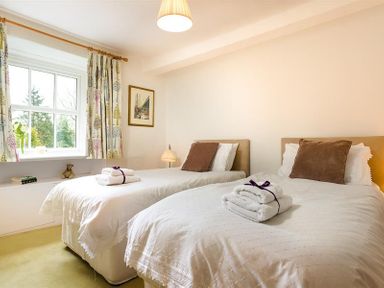 A bedroom with two single beds with white bedding brown pillows and folded towels by a window at High Cleabarrow in Windermere