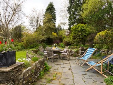 An outdoor patio with a stone floor featuring a table with six chairs and two striped deck chairs surrounded by greenery